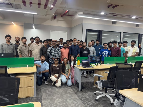 A group of approximately 26 people, primarily young men and a few women, posing for a photo in a modern office workspace with desks and computer monitors.