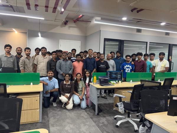 A large group of employees, mostly men, posing for a group photograph in a modern open-plan office setting with desks and computer equipment.