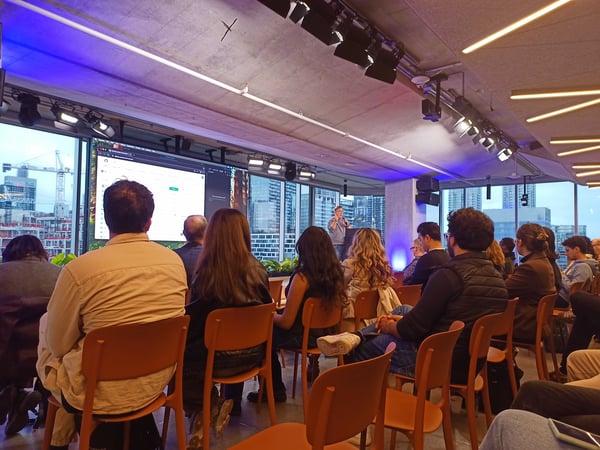 A group of people are seated in orange chairs, attending a presentation in a modern office space with large windows overlooking a city skyline at dusk.