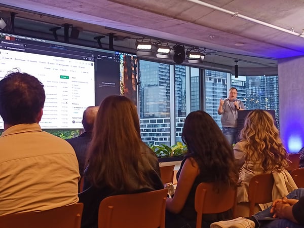 A man is giving a presentation to an audience in a modern indoor space with a large screen displaying a GitHub repository and a city view in the background.
