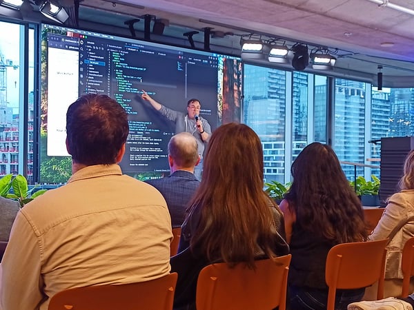 A man is giving a technical presentation to an audience in a modern office space, pointing at a large screen that displays code.