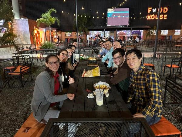 A group of people are gathered around a long wooden table at an outdoor restaurant called 'Food House' at night, illuminated by string lights.