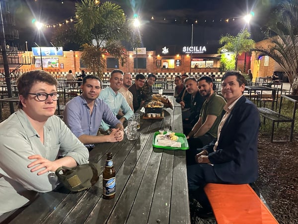 A group of people are gathered around a long wooden picnic table at an outdoor restaurant or food court at night, posing for a photo with drinks and food.