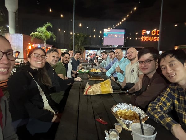 A group of about twelve friends or colleagues are gathered around a long wooden table at an outdoor food court at night, smiling for a group photo.