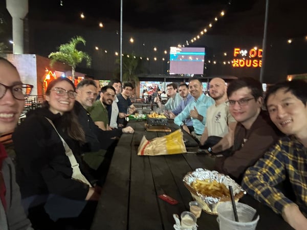A group of friends or colleagues are gathered around a long wooden table at an outdoor restaurant at night, enjoying food and drinks.