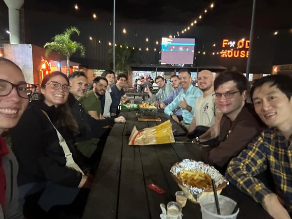 A group of people are gathered around a long wooden table at an outdoor restaurant at night, with food and drinks spread out before them.