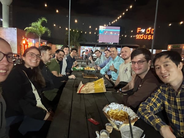 A group of friends or colleagues are gathered around a long wooden table outdoors at night, enjoying food and drinks at a venue called 'FOOD HOUSE'.