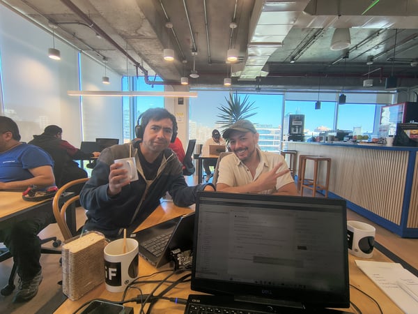 Two men pose for a photo in a modern co-working office, one holding a mug and the other making a peace sign, with laptops and a city view in the background.