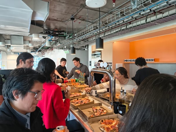A group of people are gathered in a modern office kitchen area, socializing and eating pizza from several open boxes on a counter.