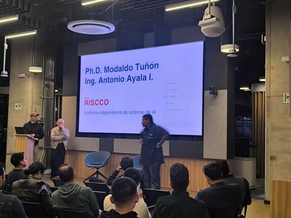 A photograph of an indoor presentation or lecture, where a speaker stands in front of a large screen displaying slides while an audience watches from their seats.