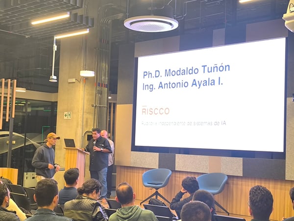 A photograph showing a presentation taking place in a modern indoor venue, with a speaker standing next to a lectern and a large screen displaying slides in Spanish to an audience.