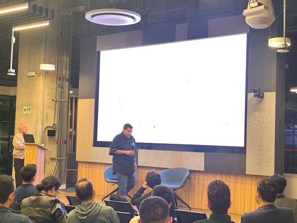 A man is giving a presentation in front of a large, bright screen in a modern conference room or auditorium, with an audience seated in the foreground.