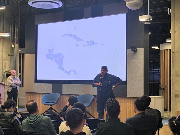 A man stands on a stage presenting to an audience, with a large screen behind him displaying a map of Central America and the Caribbean.