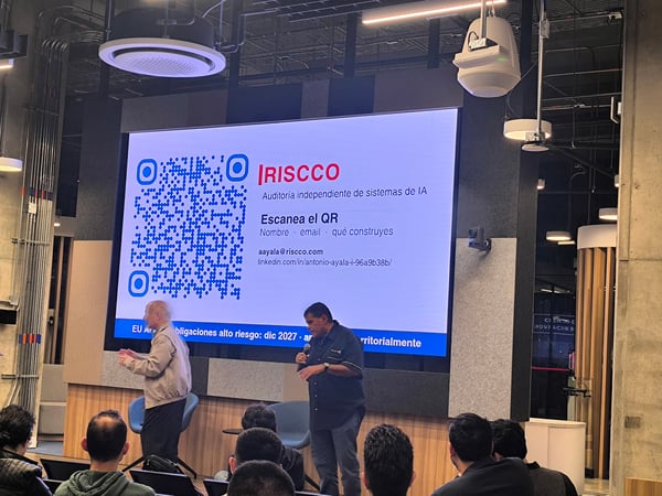 A photograph of a live presentation in a modern indoor venue, where two speakers stand on a stage in front of a large screen displaying a QR code and Spanish text, with an audience watching from the foreground.