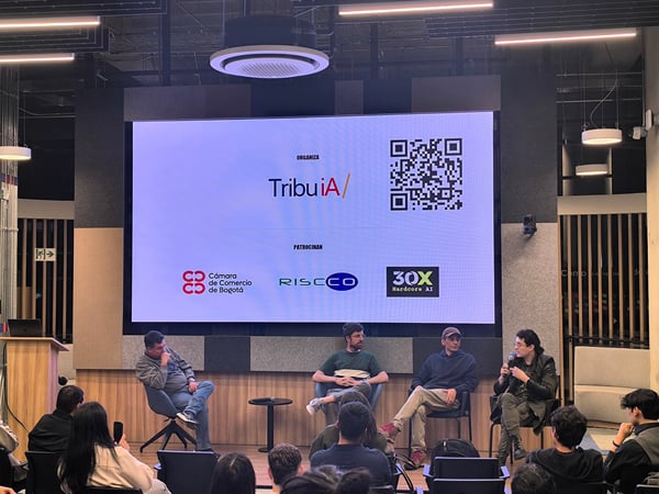 A panel discussion is taking place on a stage with four male speakers seated in front of a large screen displaying logos and a QR code, while an audience watches from the foreground.
