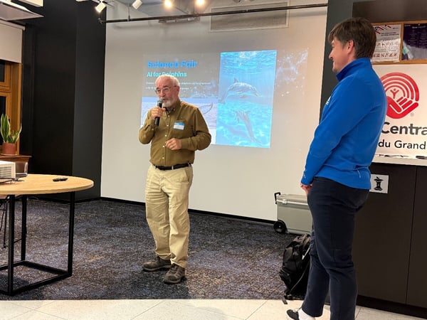 An older man with a beard is giving a presentation using a microphone in an indoor setting, standing beside a screen displaying a slide titled "AI for Dolphins" featuring images of dolphins, while another person in a blue jacket stands to the right.