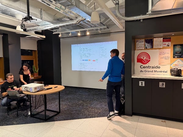 A candid photograph captures a small group of people attending a presentation or meeting in a modern office space, with content projected onto a screen on the wall.
