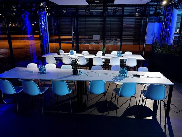An empty indoor meeting or event space featuring long white tables and numerous white chairs, bathed in dramatic blue lighting.