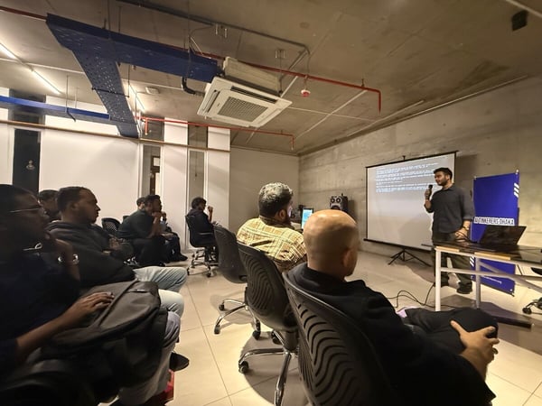 An indoor photograph captures a speaker addressing an audience during a presentation in a dimly lit room with exposed industrial ceiling infrastructure and a projection screen displaying text.
