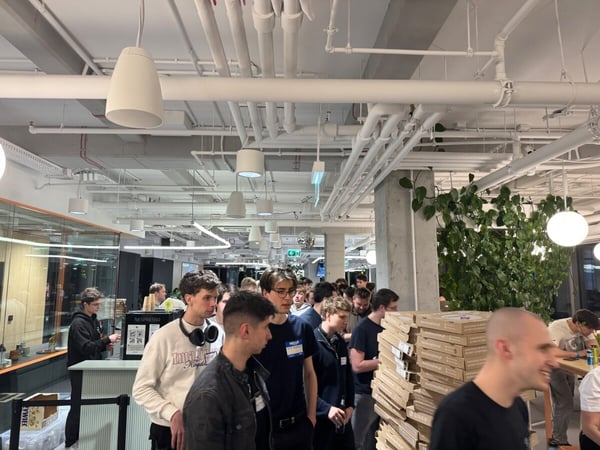 A group of young people, primarily men, are gathered in a modern office environment with exposed ceiling pipes, featuring stacks of pizza boxes and a coffee station.