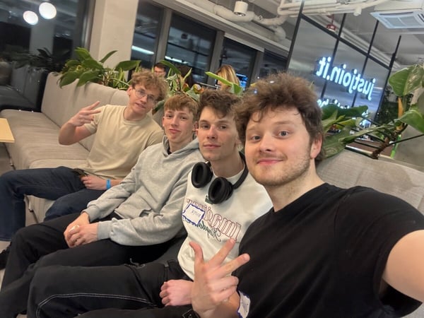 Four young men are sitting together on a long grey sofa in a modern indoor setting, likely an office or event space.