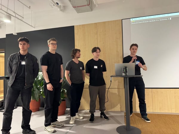 A group of five young men are giving a presentation in a modern office or event space. One man is speaking into a microphone at a high table with a laptop while the others stand beside him.