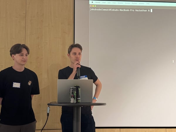 Two young men are giving a presentation at a hackathon, with one speaking into a microphone behind a laptop and energy drinks while a terminal window is projected on the screen behind them.