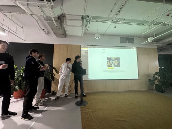 A group of young men are gathered in a modern office space for a presentation, with one person speaking into a microphone while a large screen displays a web application titled 'What's in your fridge?'.