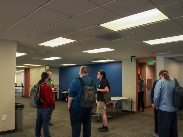 A group of people are gathered in a modern office space, engaged in a casual discussion near a blue accent wall.