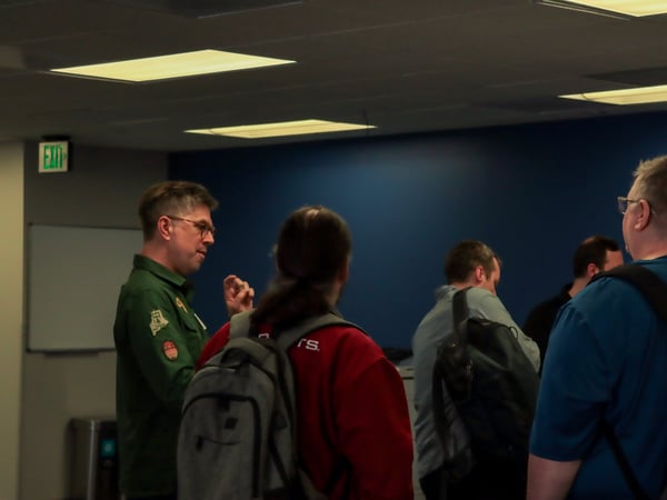 A group of men are engaged in a discussion in an indoor setting, with one man in a green shirt gesturing while others with backpacks listen.