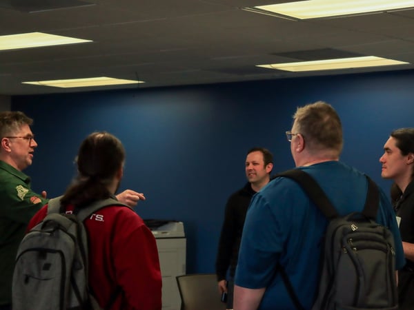A group of five people are standing in a room with a blue wall, engaged in a conversation.
