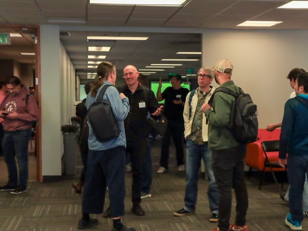 A group of people are gathered in an office hallway or event space, engaged in casual conversation while wearing backpacks.