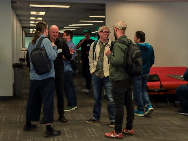 A group of people are standing and talking in an indoor hallway or office space, many wearing backpacks as if attending an event.