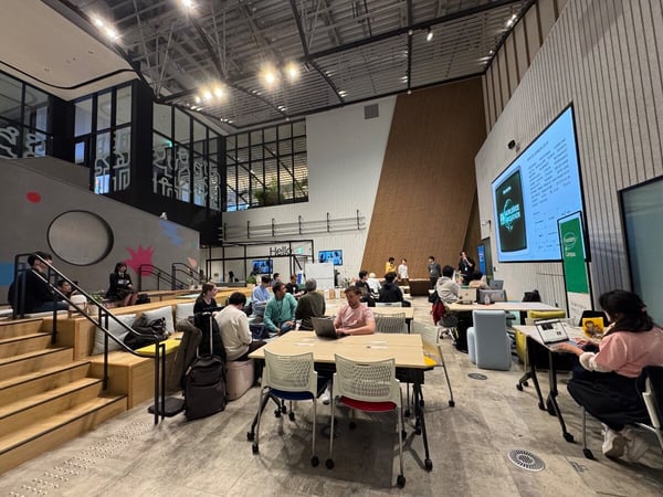 A photograph capturing a large, modern, open-plan event space or co-working area where several people are gathered, attending a presentation displayed on a large screen while others work on laptops at nearby tables.