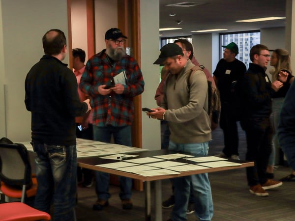 A group of people are gathered in an office environment, standing around a table covered with papers and pens.
