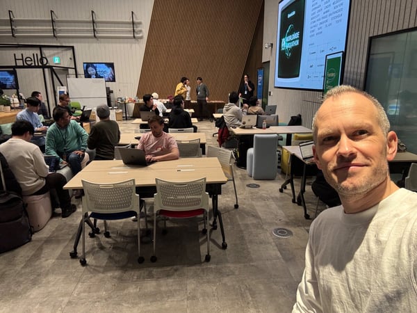 A man takes a selfie in the foreground, capturing a busy indoor event space behind him where numerous people are seated at tables working on laptops, possibly during a hackathon or workshop. A large screen in the background displays text related to a 'Worldwide Hackathon' and a list of cities.