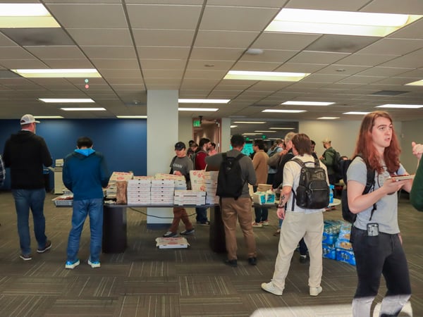A group of people are gathered in a large indoor room, likely an office or event space, around a table stacked with numerous pizza boxes.
