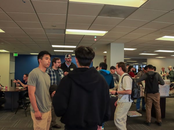 A group of people are gathered in an office-like setting for a social event, with several individuals standing and talking while pizza boxes are visible on a table in the background.