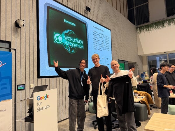 Three men stand smiling in front of a large projection screen displaying the logo and title for the "ElevenLabs WORLDWIDE HACKATHON" at an indoor tech event.