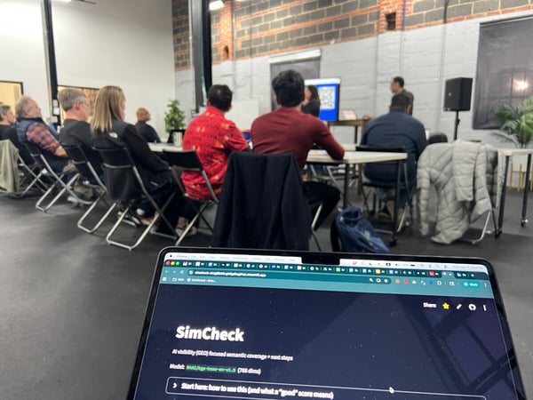 A group of people are seated in a modern workspace attending a presentation, with a laptop in the foreground displaying a web application called SimCheck.