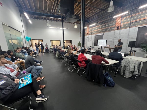 An indoor scene showing a man giving a presentation to an audience seated in a modern, industrial-style room with exposed brick walls.