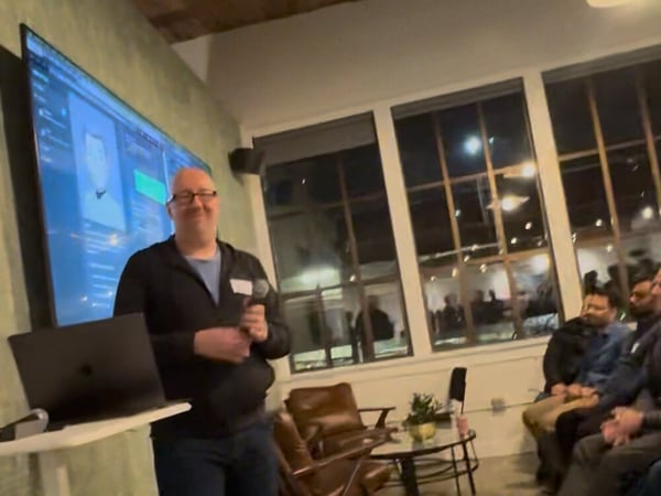 A blurry, low-light photograph shows a bald man giving a presentation or talk in an indoor venue, standing in front of a large screen displaying technical content while an audience watches from the side.