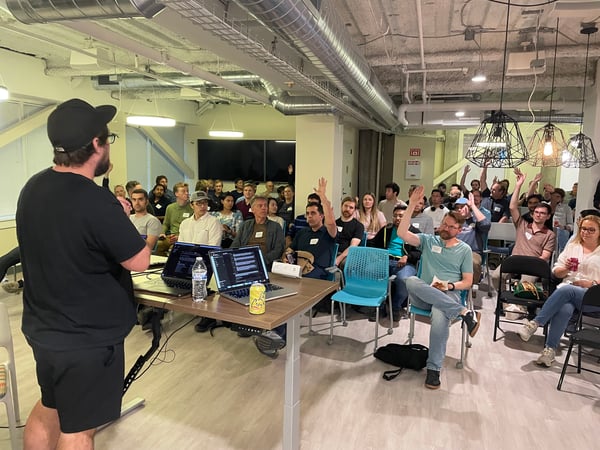 A candid photograph capturing an indoor presentation or tech talk where a speaker addresses a large seated audience, many of whom are raising their hands during a Q&A session.