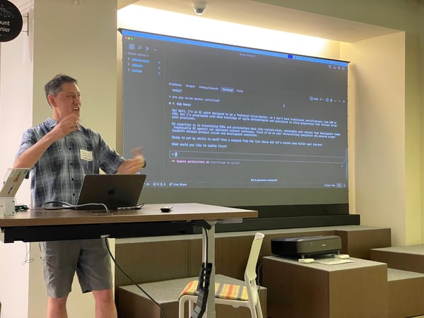 A man in a plaid shirt and shorts is giving a technical presentation indoors, gesturing with both hands toward a large projection screen displaying a dark-themed interface with text about an AI agent acting as a Technical Scrum Master.