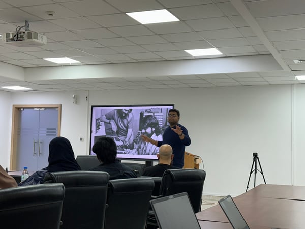 A man is standing and delivering a presentation in a conference room next to a large screen displaying a black-and-white historical photo, while several audience members watch from their seats.