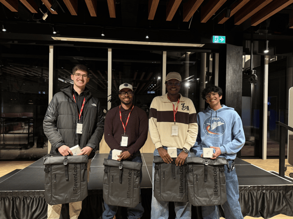 Four young men stand side-by-side on a low stage in an indoor event space, posing for a group photo while holding matching gray backpacks branded 'IIElevenLabs' and small white boxes, likely prizes or swag.