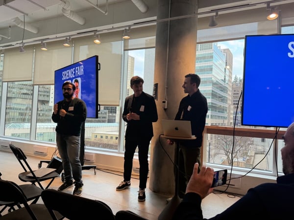 Three men are participating in a presentation or science fair event inside a modern building with large windows overlooking a city.