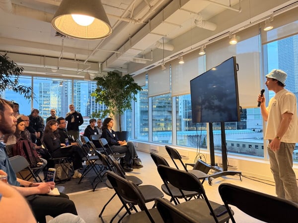 A man in a bucket hat gives a presentation to a seated audience in a modern office with large windows overlooking a city skyline.