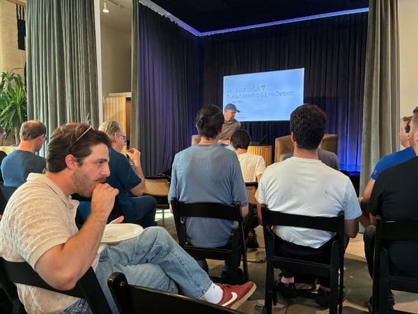A group of people sitting in chairs inside a venue, watching a presentation on a stage screen that reads 'AI Tinkerers LA Builder Meetup & Live Demos'.