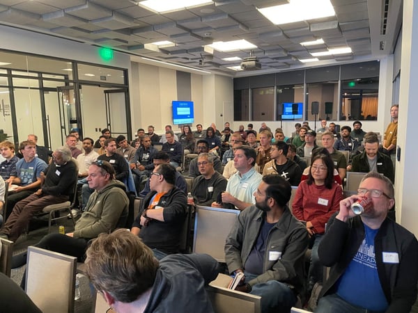 A candid photograph capturing a large audience of approximately fifty people seated in a modern conference room, attending a presentation or technical meetup.